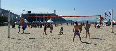 Harrastajat läiskivät palloa rannan kentillä auringonpaisteessa. Taustalla näkyvän kolossi sisällä on käynnissä olympiaturnaus.