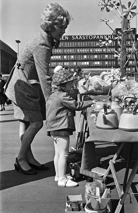 Nainen ja pieni tyttö ostamassa muovitavaraa Hakaniemen torilla, taustalla Säästöpankin talo. Kuva toukokuulta 1970.