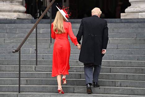 Carrie Johnson oli pukeutunut tyylikkään punaiseen pukuun ja hattuun. Boris Johnsonilla oli yllään tumma puku harmaalla liivillä ja pitkä puvuntakki.